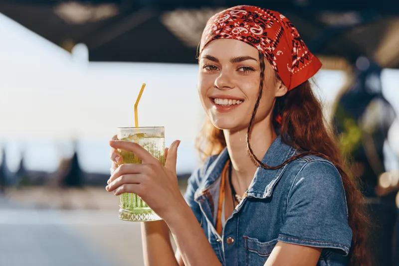Smiling woman with a red bandana enjoys a refreshing drink outdoors on a sunny day, wearing casual denim clothing and radiating happiness and relaxation.