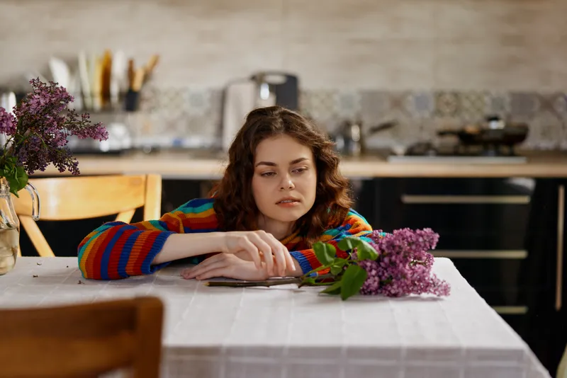 Woman in striped sweater sits at a dining table alone at home, resting her head on her arms, with wilted flowers nearby, conveying contemplative mood and quiet domestic scene