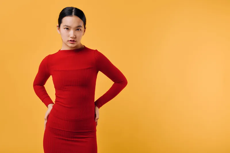 Woman in red dress standing with hands on hips against orange background for fashion, confidence, style and beauty portrait. Female model posing with serious expression and modern look.