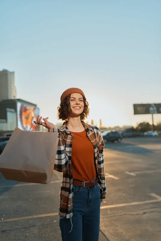 Woman shopping with a smile holding a paper bag in an urban parking lot, candid lifestyle portrait with authenticity and golden hour glow, mindful living and emotional storytelling.