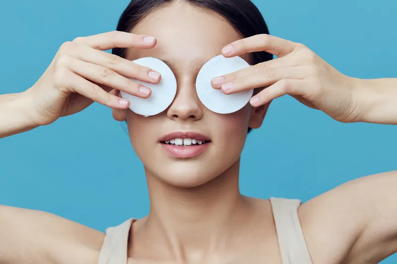 Young woman holding cotton pads over her eyes, promoting skincare and relaxation with a serene expression against a vibrant blue background