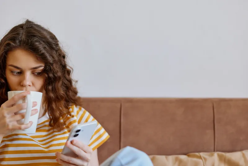 Casual woman indoors on a couch, sipping from a mug while scrolling a smartphone, cozy home scene with warm light and relaxed mood, striped shirt, comfortable ambiance