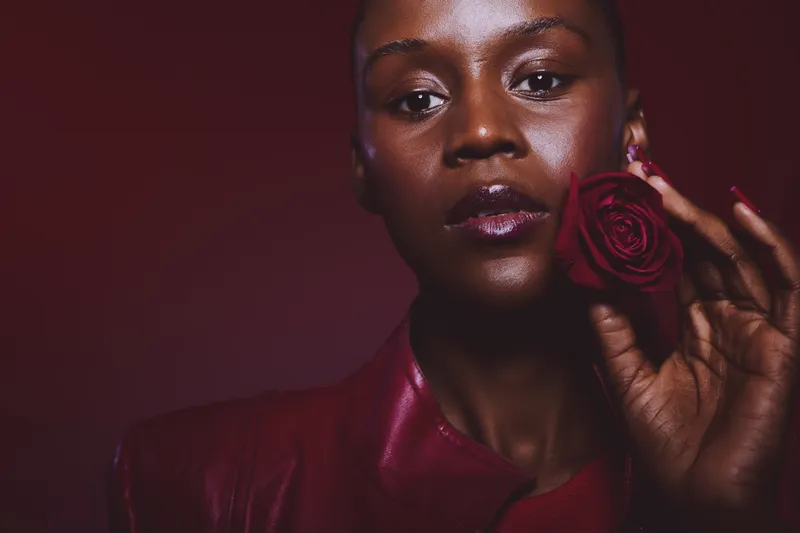 Close-up portrait of a dark skinned African woman holding a red rose near her face. She wears a dark red leather jacket against a deep red background. Concept of beauty and elegance.