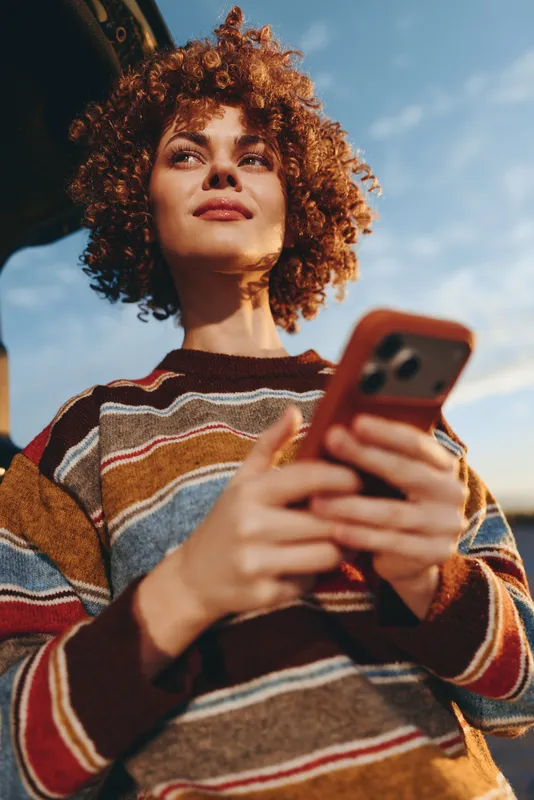Woman lifestyle selfie smartphone outdoors, curly hair, wearing striped sweater, holding phone toward camera, sunny sky background, relaxed confident mood, summer travel vibe, outfit