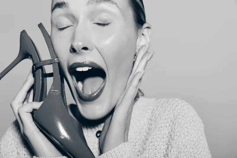 Excited Caucasian woman with fair skin holds red patent leather shoe near face, eyes closed and mouth open, expressing joy and surprise.
