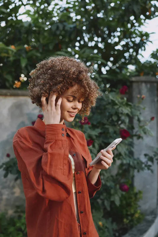 A woman in a rust cardigan checks her smartphone outdoors, smiling softly in a sunny garden, a candid lifestyle moment