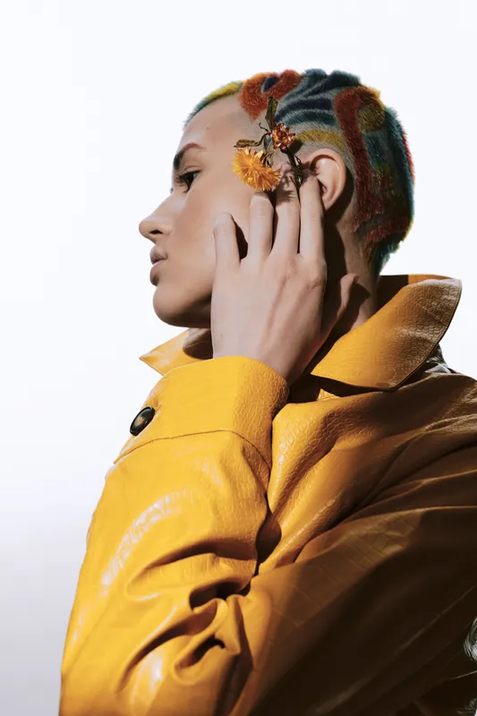 Woman with colorful buzzcut hairstyle and yellow jacket posing with dried flowers near ear, showcasing unique fashion style against plain white background.