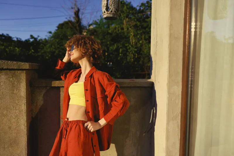 A stylish woman in a red jacket and yellow crop top stands on a sunlit balcony, looking away with a hand on her head. Outdoor lifestyle fashion moment
