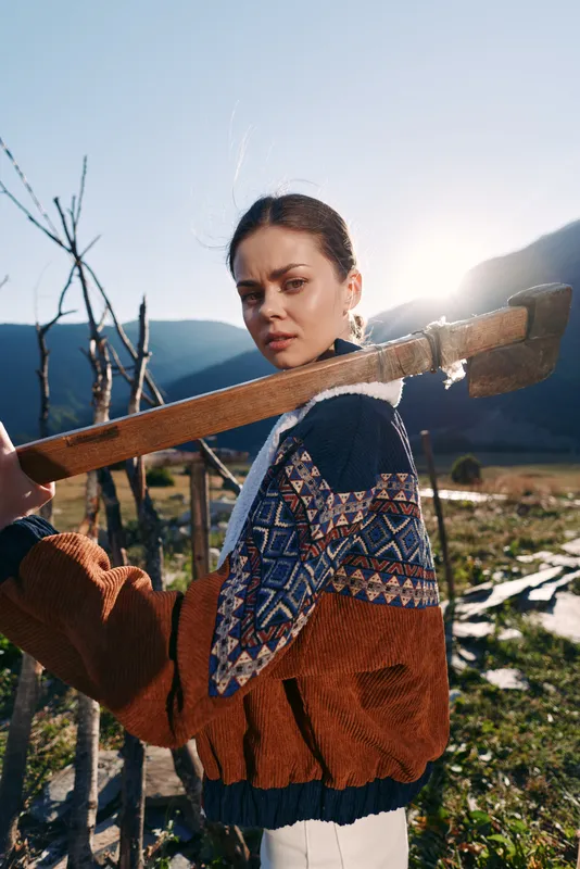 Woman portrait with axe over shoulder in rural mountains at sunrise, sunlight backlight and rustic jacket. Outdoors farmer model with confident gaze, traditional knit sweater and fence scene.