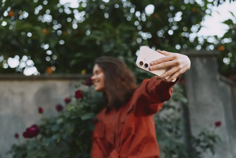 A woman in a red blouse takes a playful selfie in a sunlit garden, with flowers and greenery in the background. A lifestyle portrait capturing casual mood and personal expression