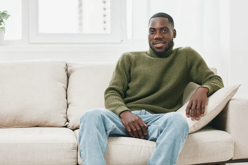 Relaxed young Black man smiling in a cozy living room, wearing a green sweater and blue jeans, representing comfort and happiness at home