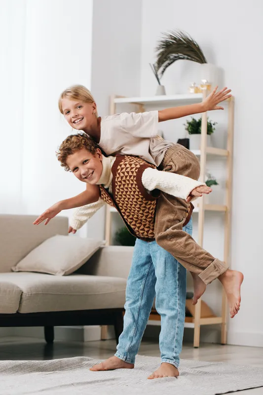 Playful moment captures siblings sharing a joyful piggyback ride in a bright living room, smiles, relaxed mood, casual outfits, soft natural light, cozy home setting, spontaneous carefree fun and