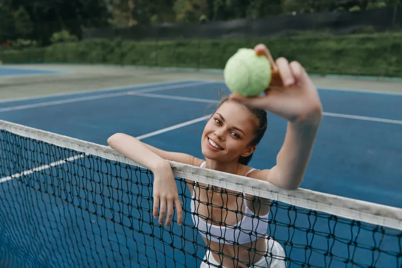 Happy young female tennis player in sportswear holding a tennis ball at the net on a sunny day, showcasing energy and excitement on a blue court