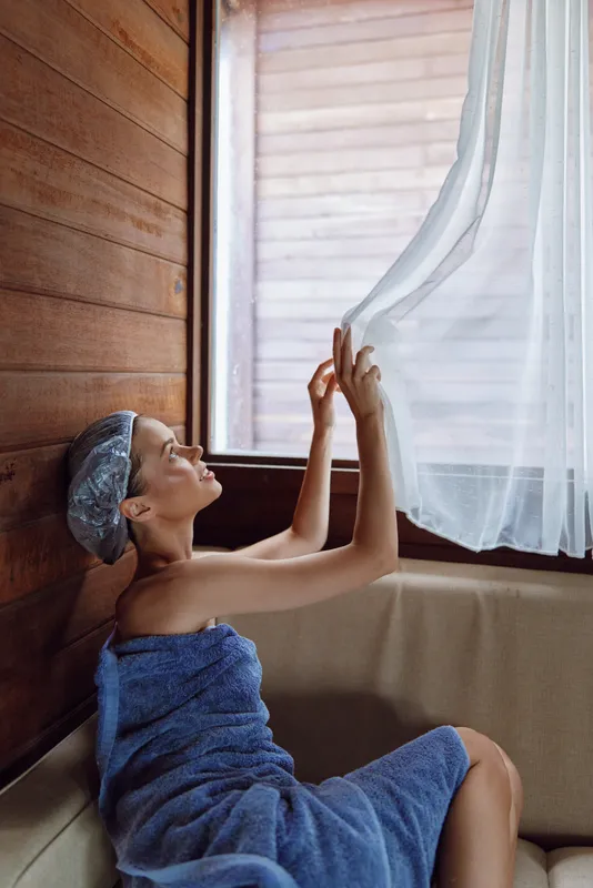 Relaxed woman wrapped in blue towel enjoying morning light while gently touching sheer curtain in cozy wooden room, peaceful moment near window.