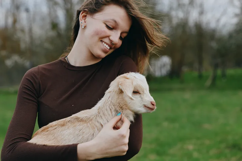 Goat, kid, woman, outdoor, lifestyle, nature, countryside, portrait, smiling, affection in a sunlit moment as a gentle breeze moves through greenery and calm happiness in a pastoral setting
