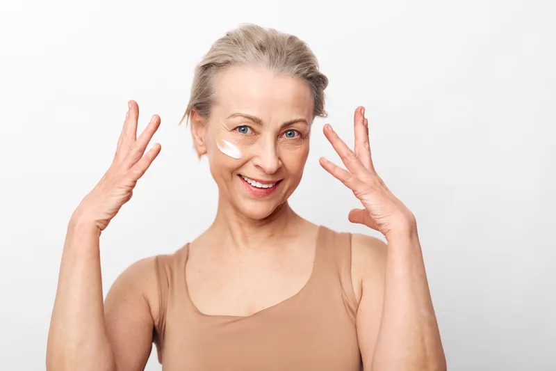 Mature woman with short hair applies moisturizing cream on face smiling, isolated on white background. Concept of skincare, healthy aging, natural beauty, and self-care routine for women.