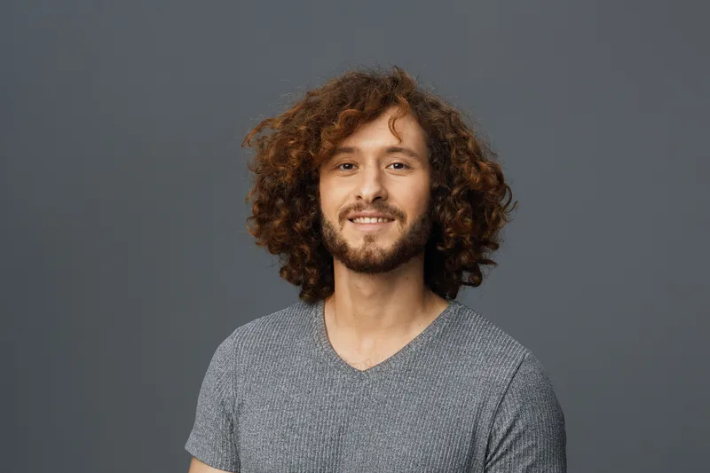 Smiling young man with curly hair in gray shirt, showcasing confidence and positivity against a minimalist gray background, perfect for lifestyle and wellness concepts