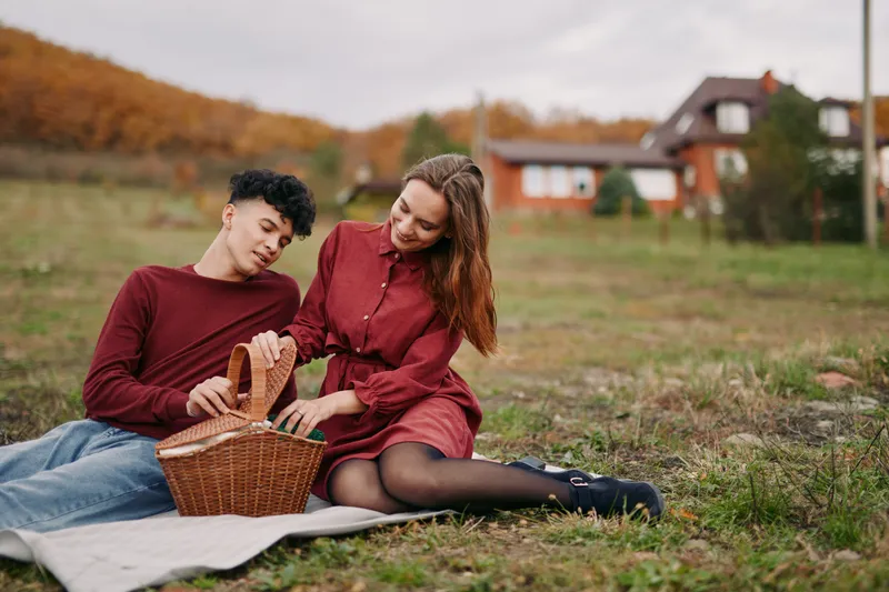 A candid moment between a couple sits on a blanket in an autumn field, a wicker basket beside them, warm light highlighting genuine emotion and simple, everyday connection.