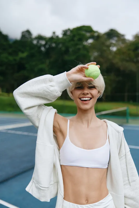 Cheerful young woman in sports attire holding a tennis ball, showcasing a healthy lifestyle on a tennis court surrounded by lush greenery