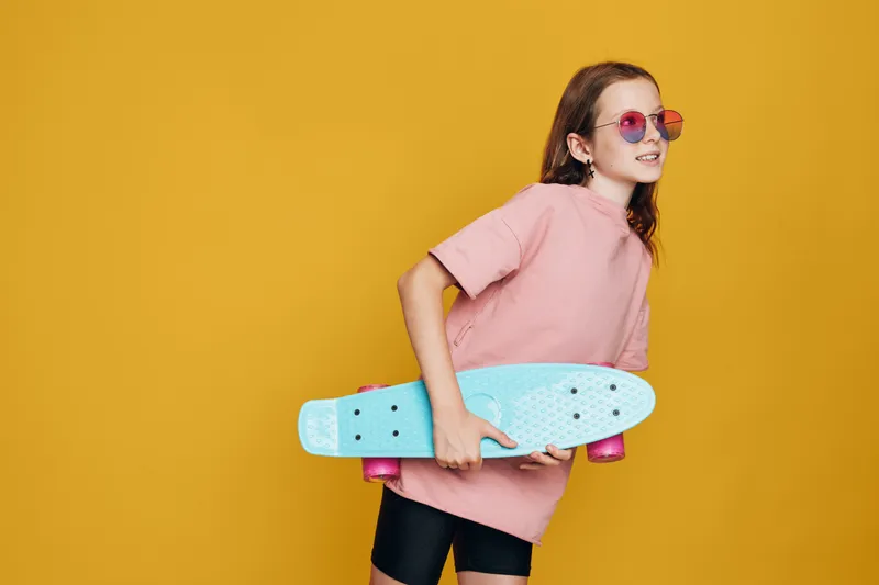 Young girl in pink t-shirt holding a blue skateboard with pink wheels, wearing sunglasses and smiling. Studio shot on a yellow background. Concept for youth, sport, and lifestyle.