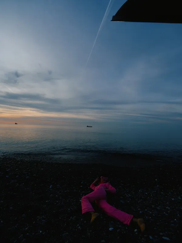 Beach scene at dusk with a person in pink clothing lying on the pebbled shore, calm sea, distant boat, and a dark wing overhead creating a dramatic silhouette.