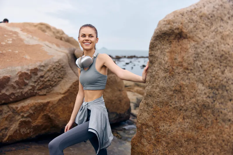 Young woman enjoying a workout on the beach, wearing athletic clothes and headphones, smiling in a natural rocky setting