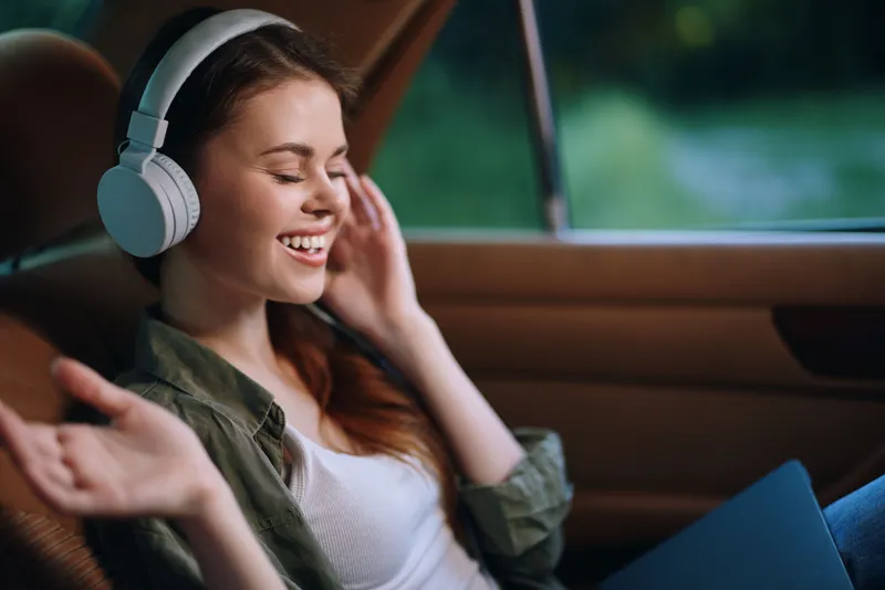 Young woman enjoying music in a car with headphones, relaxed and cheerful expression, natural background, comfortable setting, afternoon light.