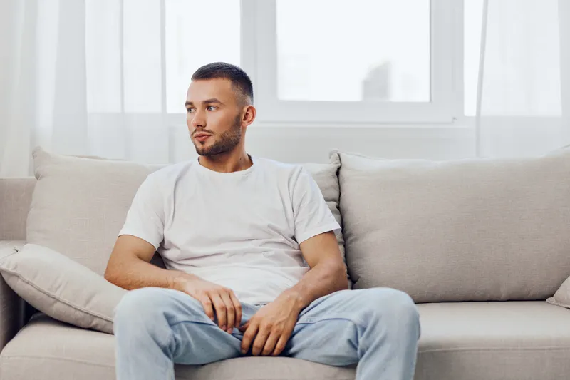 Thoughtful man relaxing on a cozy couch, casual outfit, modern interior, soft color palette, perfect for lifestyle and relaxation themes.