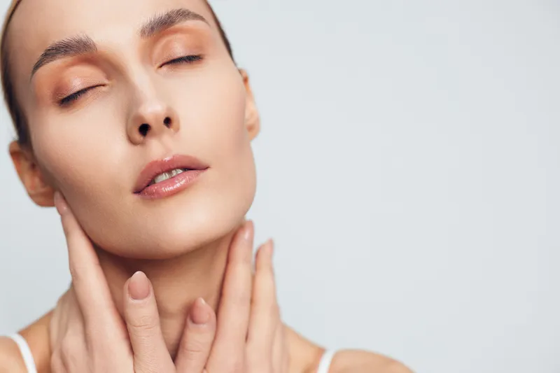 Young woman with closed eyes and natural makeup gently touches her neck with both hands isolated on light background, showing calm and relaxed facial expression and smooth skin.