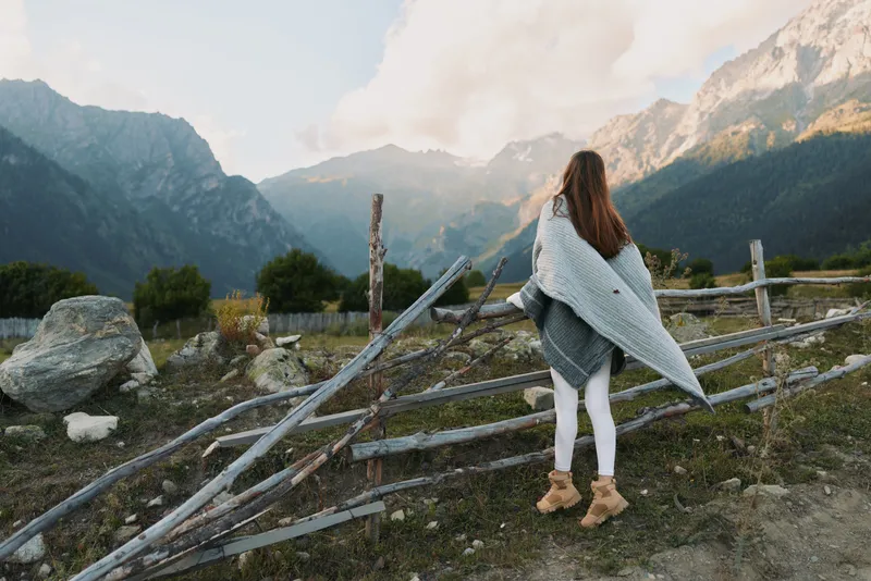 Young woman wrapped in a gray blanket stands near a rustic wooden fence looking at mountain peaks under a cloudy sky during autumn.