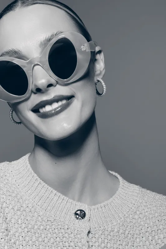 Smiling white woman with pale skin wearing large sunglasses and hoop earrings poses in knitted sweater. Studio portrait with soft lighting for fashion, accessories, and beauty concept.