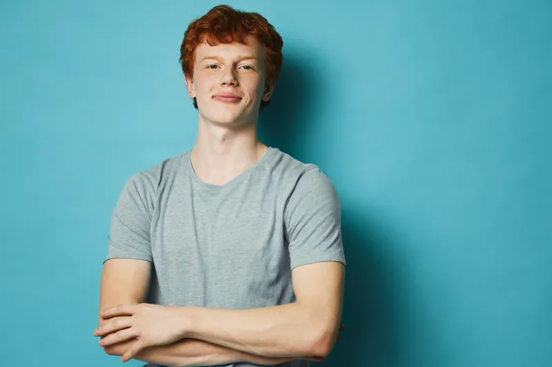 Redhead young man in gray t shirt standing with arms crossed smiling confident on blue background. Casual portrait, studio lighting, relaxed pose, friendly and approachable expression.