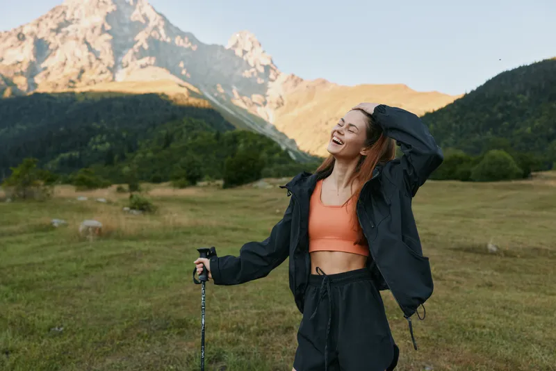 A cheerful young woman enjoys the outdoors, hiking on a grassy trail with mountain peaks in the background. She smiles and radiates energy during her scenic adventure.