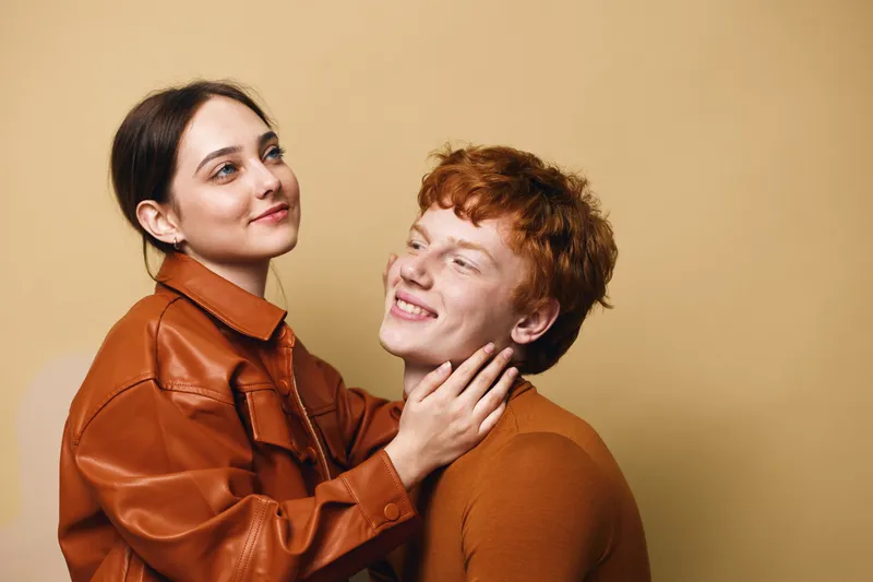 Couple with loving expression posing together indoors with beige background. Woman in brown jacket gently holding smiling man in sweater, showing affection and connection.
