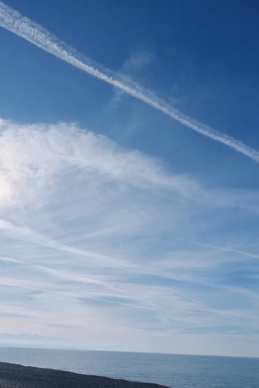 Blue sky over calm sea and pebbled beach with wispy clouds and contrails, a tranquil coastal seascape at the horizon.