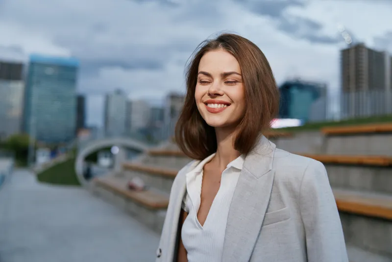 Confident businesswoman smiling outdoors in a city setting, dressed in a professional outfit, exuding positivity and success amidst an urban backdrop.
