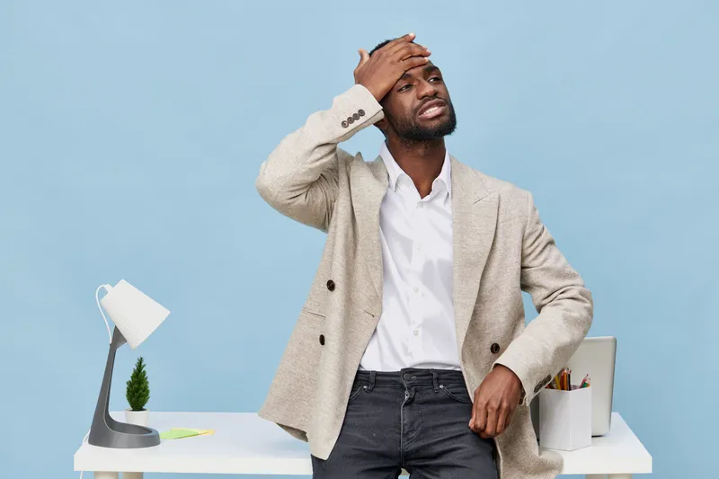Exhausted stressed young African American man in formal wear with blazer and white shirt, holding his head, standing near modern white desk and lamp, feeling overwhelmed, indoor office vibe, concept o