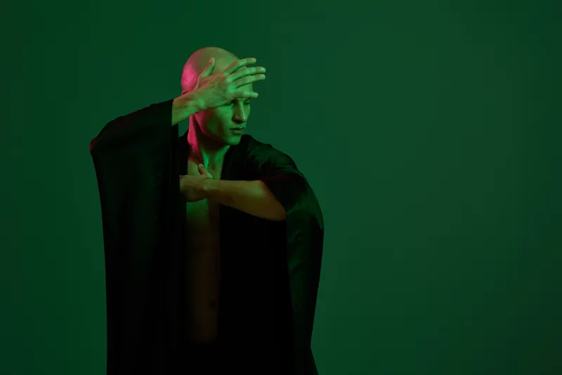 A man in a dramatic pose wearing a flowing black robe, illuminated by vibrant green and pink lights. This image captures a striking and artistic moment.