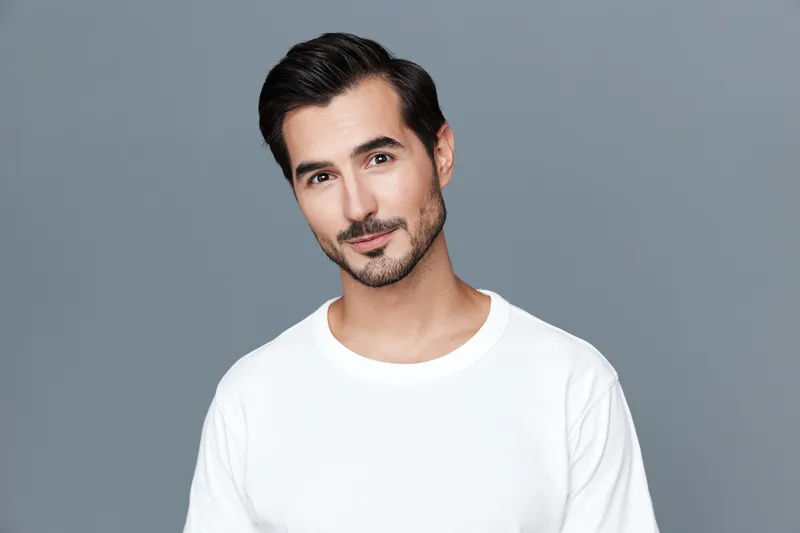 Handsome man with dark hair wearing a stylish white t-shirt, looking confidently at the camera against a gray background during a studio portrait session.
