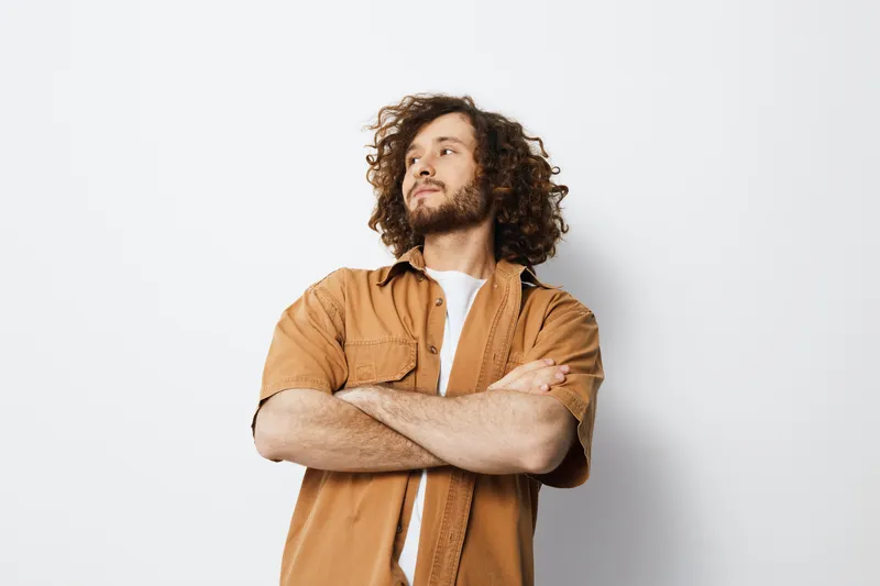 Confident young man with curly hair in a casual brown shirt, standing against a white background, showcasing a thoughtful expression and relaxed pose