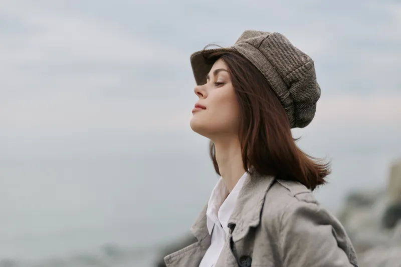 Young woman in vintage hat and coat stands outdoors with closed eyes, enjoying fresh air near sea. Calm mood, fashion, and autumn concept in natural light over cloudy sky.