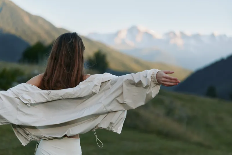 A lone person stands in a expansive meadow with arms outstretched, facing distant mountains under a clear sky, evoking freedom, serenity, and expansive possibility.