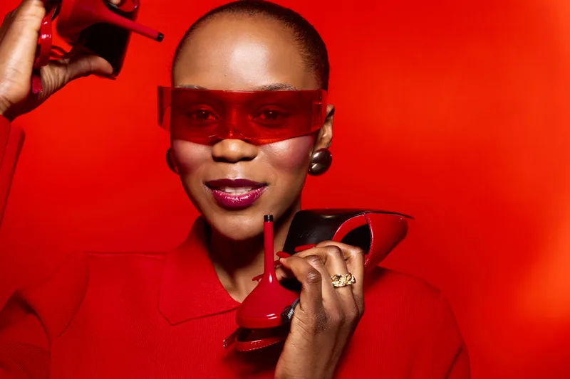 Stylish black woman with dark skin wearing red sunglasses and red outfit holds red high heel shoe and lipstick, posing against red background. Concept of fashion, beauty, and confidence.