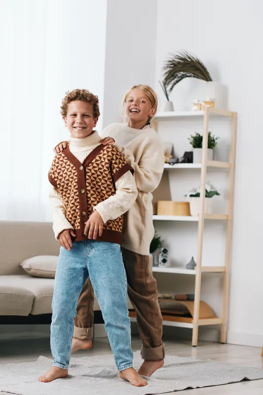 Two cheerful children play indoors, smiling and hugging in a bright living room bathed in sunlight. Cozy sweaters, denim jeans, soft textures, and a warm, carefree vibe.