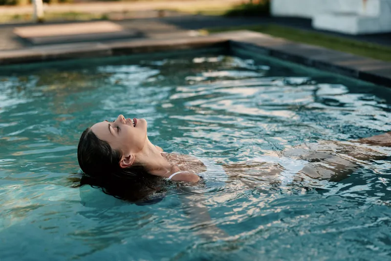 Relaxed woman floating in a pool with dark hair, enjoying a calm moment. Summer leisure, wellness, and self care vibe. Soft light reflects on water for a serene mood.