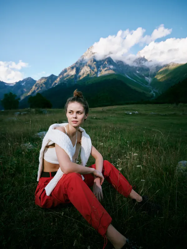 Woman sitting on grass in alpine landscape with mountains wearing red pants and white sweater outdoor fashion and serene nature scene with blue sky and peaceful mood