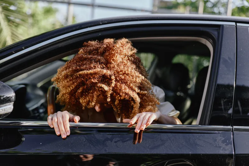 African American woman with curly hair leaning out of a black car window, bright daylight, casual white shirt, outdoor urban setting, natural light.