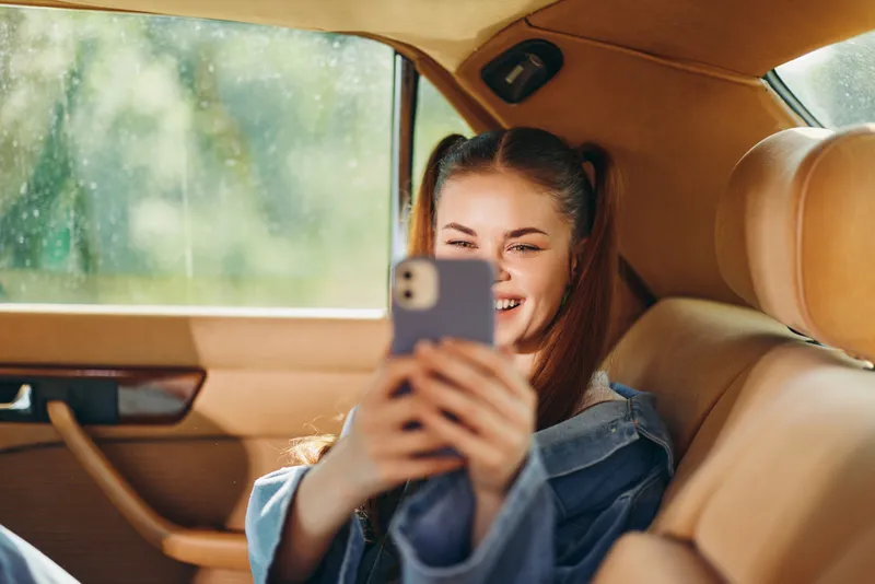 Happy teenage girl with pigtails smiling and using smartphone in a vintage car, capturing a carefree moment in a sunny setting.