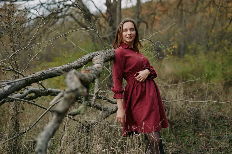 A genuine portrait of a pregnant woman in a red dress leaning on a fallen tree in a natural meadow, conveying authenticity and calm confidence with a realistic maternal connection and presence.