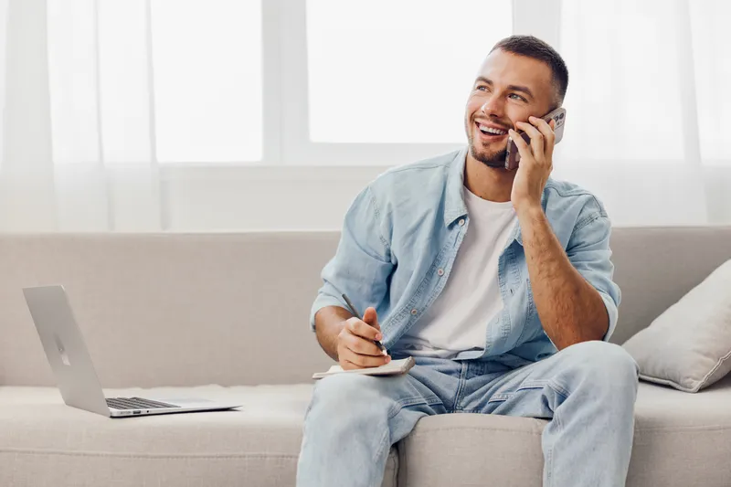 Smiling man engaging in a phone call while taking notes, reflecting emotional minimalism and soft masculinity in a relaxed home setting.